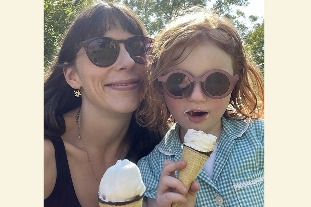 On the left, a lady with long dark hair, earing sunglasses, smiling and holding an ice cream, on the right a young girl with wavy hair, in a blue and white checked dress, also wearing sunglasses and holding an ice cream.