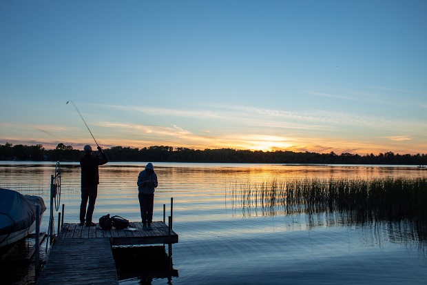 Two people fishing on a dock, with a sunset in the distance.