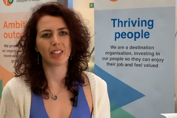 A lady with dark curly, shoulder length, hair standing in front of a sign that says ‘thriving people’.