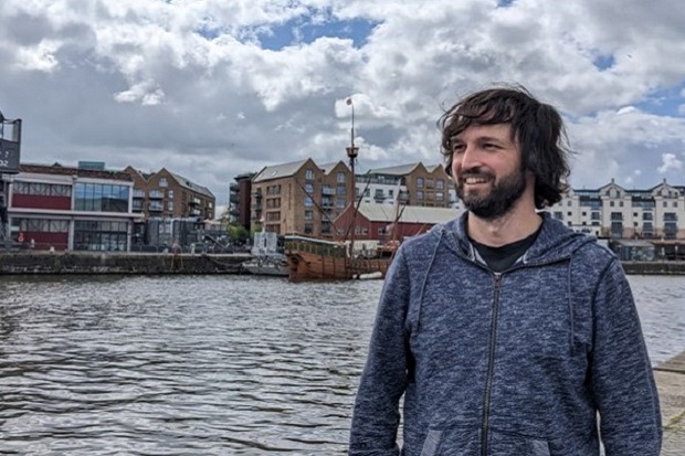 A man with dark hair and beard standing by a harbour.