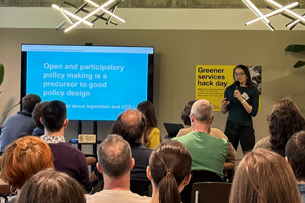 A person standing in front of a large screen looking at an audience. The screen says: ‘Open and participatory policy making is a precursor to good design’.