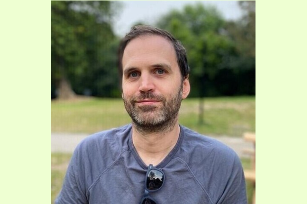 A man with short dark hair and a beard wearing a grey t shirt with sunglasses draped over the front of the t shirt.