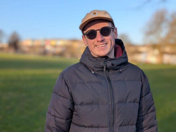 Jason is stood in a park. He is in focus in the foreground, with grass, trees and a blue sky behind him. Jason is wearing a cap, sunglasses and a blue jacket.