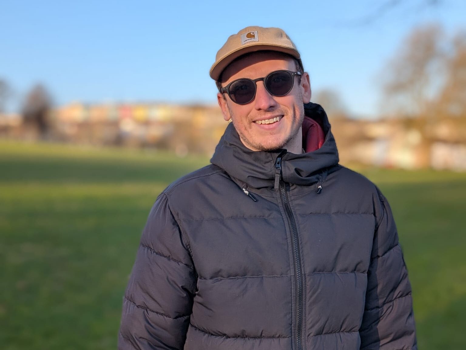 Jason is stood in a park. He is in focus in the foreground, with grass, trees and a blue sky behind him. Jason is wearing a cap, sunglasses and a blue jacket.