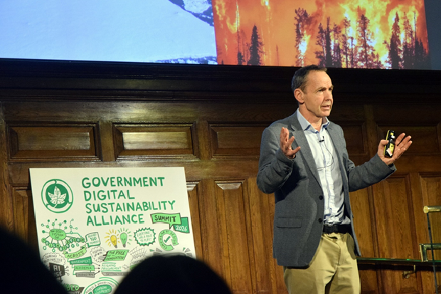 Photograph of a speaker presenting at the Government Digital Sustainability Alliance Summit 2026, with a wooden panelled background and a large illustrated poster highlighting sustainability themes such as renewable energy and environmental protection. The speaker gestures with hands while holding a remote, and a projected image of a wildfire contrasts with a glacier, emphasizing climate change challenges.