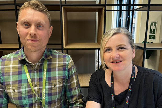 Photograph of two people sitting indoors in front of empty wooden shelves, both wearing visitor lanyards. One person wears a green and purple plaid shirt with a yellow lanyard, and the other wears a black shirt with a black lanyard featuring colourful logos.