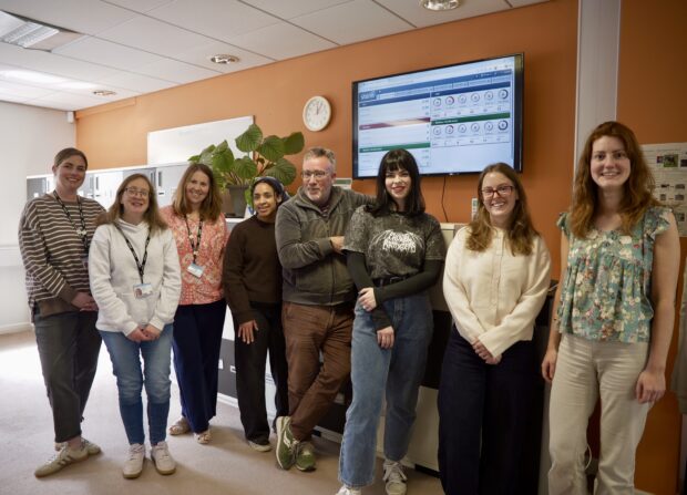 Eight people from the Customer Advice Team and Improving Customer Experience team stand together in an office workspace against an orange wall. Behind the group are filing cabinets, a large potted plant, a wall clock, and a wall-mounted monitor displaying a performance dashboard. The people are arranged in a row facing the camera, wearing a mix of casual and business-casual clothing.