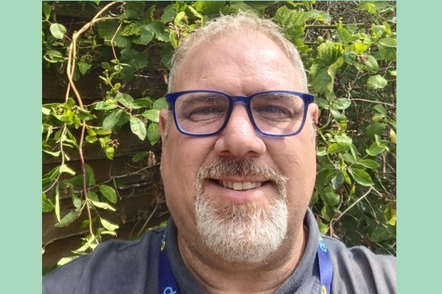 Photograph of a middle-aged man with glasses and a gray beard smiling outdoors in front of green leafy plants. The close-up portrait highlights his friendly expression and casual attire with a lanyard around his neck.