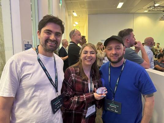 Photograph of three smiling people wearing conference badges standing indoors at a networking event. Two men and one woman are in the foreground, with the woman holding a small award or trophy, while several other attendees are visible in the background.