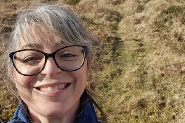 Photograph of a smiling woman with glasses and gray hair standing outdoors on a grassy, sunlit hillside. The image captures a casual, cheerful moment with natural background featuring dry grass and a clear sky.