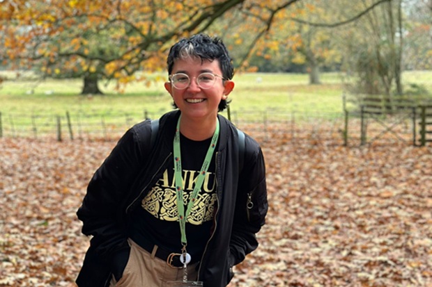 Photograph of a smiling person with glasses, short hair, black jacket, graphic shirt, and green lanyard standing outdoors in an autumn park. Fallen leaves, orange foliage, grassy field, tree-lined background, and rustic fencing create a seasonal setting.