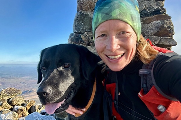Photograph shows smiling hiker in green headscarf posing with black dog beside a rocky summit cairn. Bright blue sky, expansive mountain landscape, hiking gear, and dog’s grey muzzle emphasise outdoor adventure and companionship.